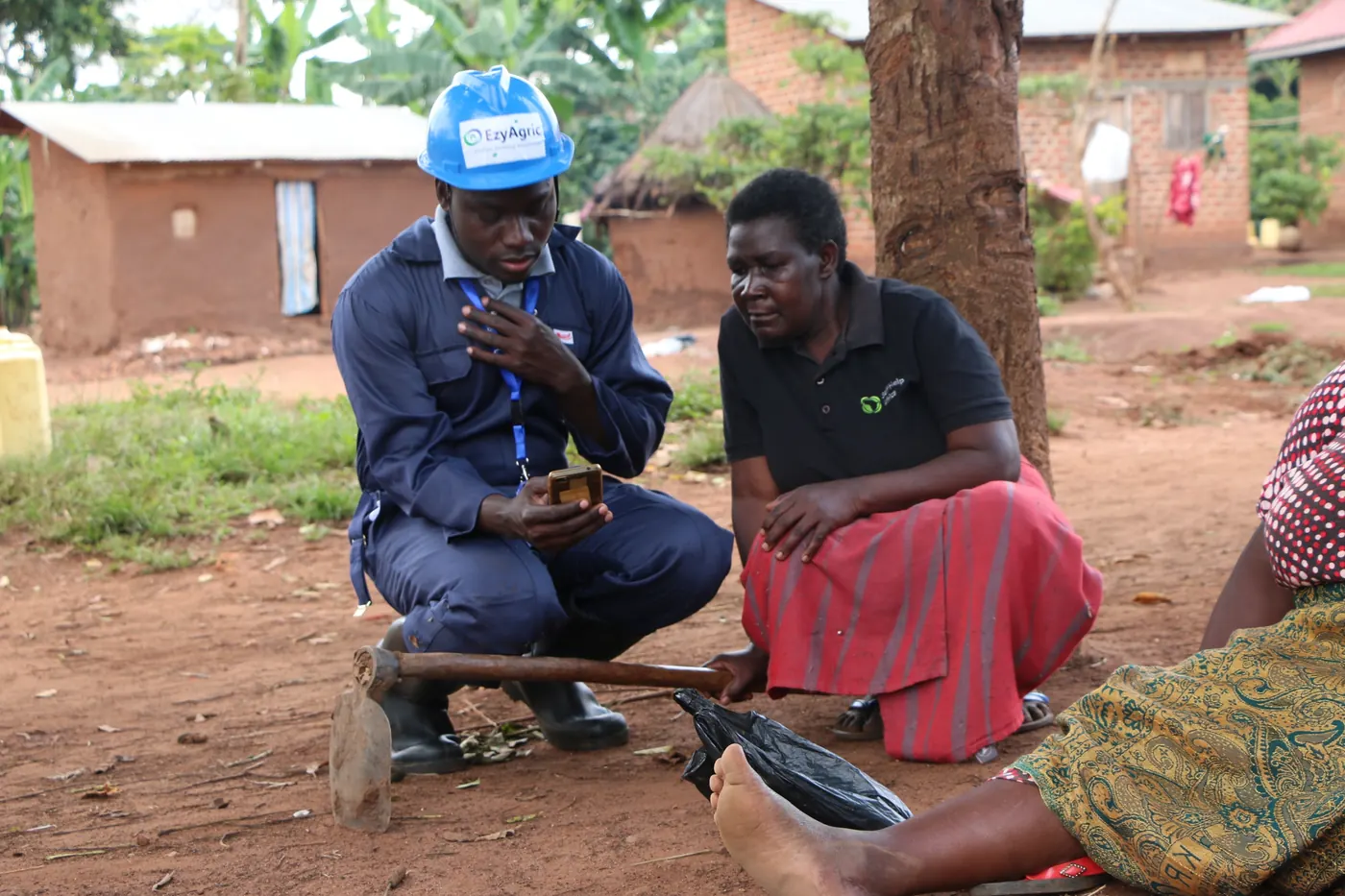 An EzyAgric agent in a branded helmet shows a farmer something on his phone, outside her home