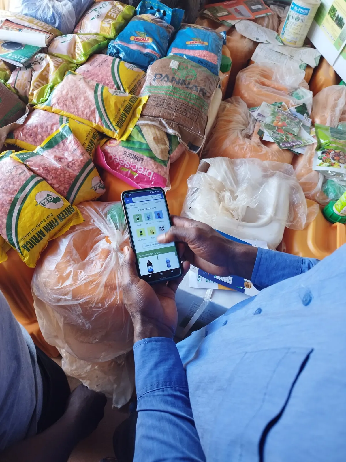 Someone browses Agrishop's "Shop by category" page on a phone, over sacks of maize seed and bottles of crop protection products