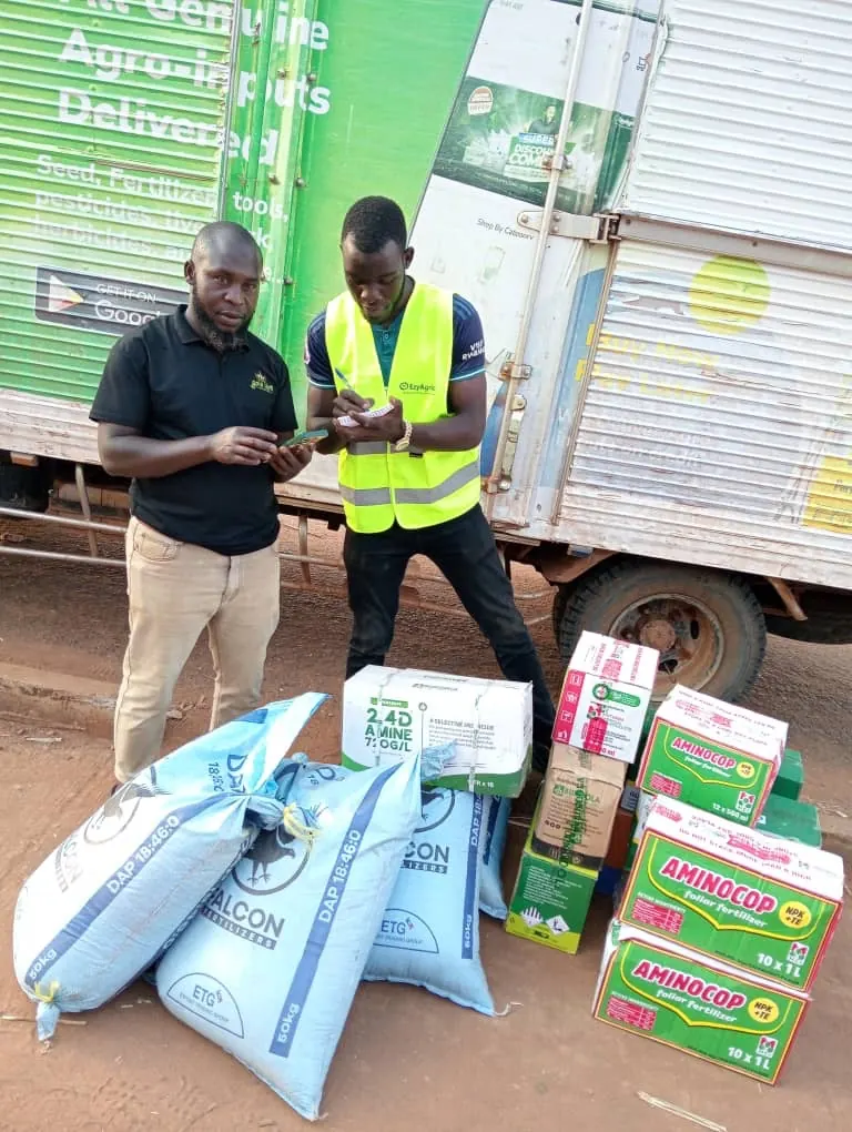 An EzyAgric delivery agent in a high-visibility vest writes up a delivery beside the truck while the customer checks his phone, with sacks of DAP fertilizer and boxes of herbicide and foliar fertilizer at their feet