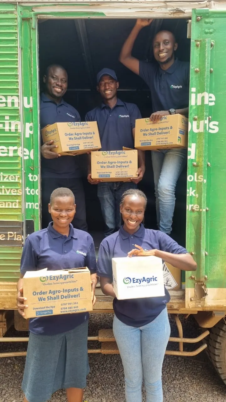 The EzyAgric delivery team, three in the back of the truck and two in front, smile with boxes reading "Order Agro-Inputs & We Shall Deliver!"