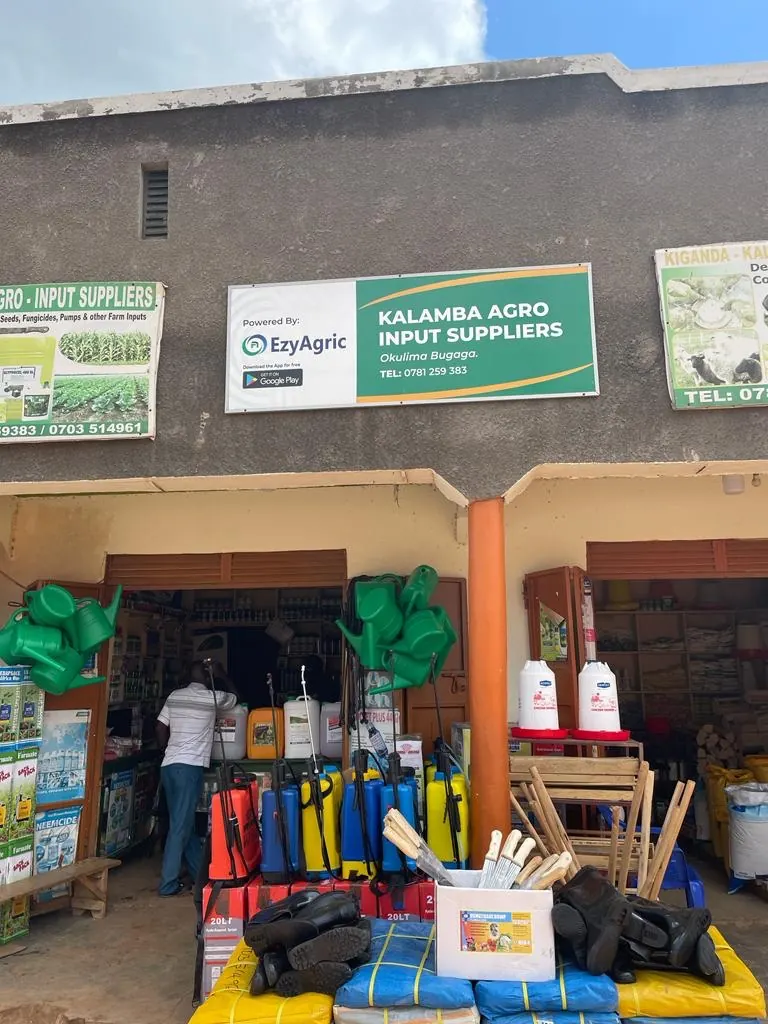 Kalamba Agro Input Suppliers, an agro-input shop with a "Powered by EzyAgric" sign, with sprayers, watering cans, boots and sacks of inputs out front