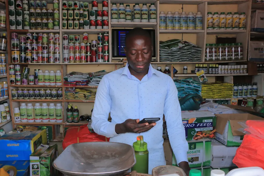 An agro-input dealer at his counter with his phone, shelves of seed and crop protection products behind him