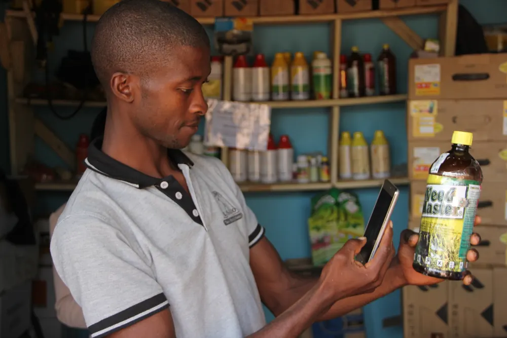 A young farmer in an agro-input shop checks a bottle of herbicide against his phone